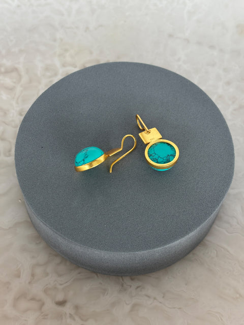 Angled Shot of Turquoise Gem Dangle Kaira Earrings Made With Sterling Silver and Gold Plated on Grey Pillow